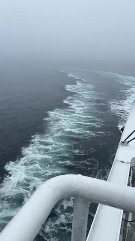 The video shows a view from a moving boat or ship, looking out at a dark, choppy sea and a foggy horizon. The wake of the boat is visible on the right side of the frame, with white foam and churning water. In the distance, a small white boat can be seen through the fog. The railing of the boat from which the video is filmed is visible in the foreground. The overall atmosphere is overcast and moody.