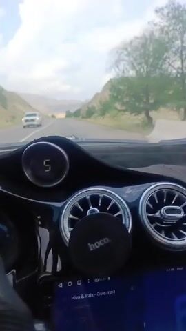 The video shows the view from the driver's seat of a car driving on a road through a valley. The dashboard of the car is visible in the foreground, with a digital display showing "5" and "0" at different times, along with air vents and a phone mount. The road ahead is paved and appears to be in good condition. On either side of the road are hills with sparse vegetation and trees. The sky is cloudy with patches of blue. Another vehicle is visible on the road in the distance on the left side. The overall scene suggests a scenic drive through a rural or mountainous area.