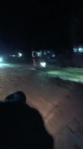 The video shows a nighttime view from the perspective of someone riding a motorcycle. The rider's shadow is visible in the foreground, cast by the motorcycle's headlight. The road is unpaved and appears to be dusty or muddy. Several buildings and streetlights are visible in the distance, along with the headlights of other vehicles. In one instance, another motorcycle with a rider passes by. The video is shaky, indicating movement.