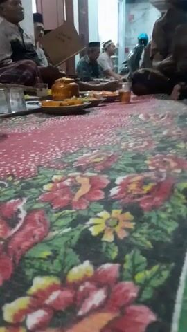 The video shows a group of men sitting on the floor, which is covered with a colorful patterned rug. They are in what appears to be a home or community gathering. Some of the men are wearing Islamic caps. In the foreground, there are plates of food and cups of tea. Some of the men are holding and looking at books or papers, possibly religious texts. One man in the background is holding a book. The overall atmosphere seems to be one of a shared meal and religious or communal discussion.