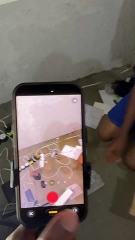 A person wearing blue shorts and a grey t-shirt is kneeling on a concrete floor next to a white wall. The person is opening a plastic bag with an orange cap. A cell phone in a holder is recording the person. On the floor around the person are papers, wires, tools, and other items.