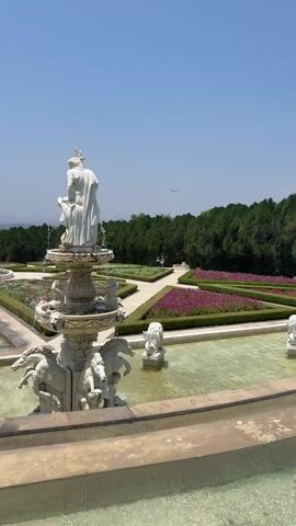 The video shows a formal garden with a large white fountain in the foreground. The fountain has multiple tiers and is adorned with statues, including horses and a human figure at the top. Water is flowing from the fountain. Behind the fountain is a patterned garden with trimmed hedges and flowerbeds in shades of purple and white. A pathway runs through the garden, and two people are seen walking in the distance. The background features a row of tall, green trees. The sky is blue and mostly clear. An airplane flies overhead in the distance.