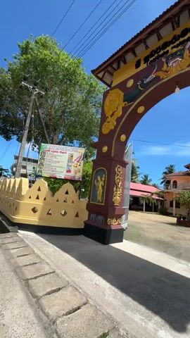 The video shows a bright, sunny daytime scene, focusing on an ornate, arch-shaped entrance gate. The archway is predominantly dark red and yellow, adorned with intricate golden carvings of mythological creatures and human figures. In Sinhalese script, "ශ්‍රී සප්තපරාජය - 1925" (Sri Sapthaparajaya - 1925) is inscribed on the arch. To the left of the archway, a low wall with yellow and dark brown details is visible, along with a utility pole and a banner displaying Sinhalese text and images. Beyond the arch, a spacious, unpaved courtyard leads to a multi-story building with traditional Sri Lankan architecture, featuring a distinctive roof and balconies. The sky is clear and blue with sparse white clouds. The camera moves forward, entering the premises through the archway, revealing the building and surrounding palm trees. The location is Walgama, Sri Lanka.
