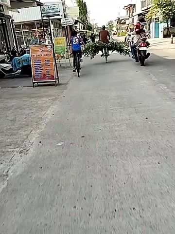 A street-level shot captures a sunny day in what appears to be an urban or semi-urban setting, with buildings lining the road. Two individuals on bicycles are visible: one, a child, cycles in the foreground wearing a blue and purple shirt with the number "29" on the back. The other, an adult, rides a bicycle with a large bundle of green branches secured to the back, extending horizontally. As the video progresses, the child on the bicycle turns to face the camera briefly before continuing to cycle. A third child on a smaller red bicycle briefly appears between the first child and the adult. In the background, a person on a scooter or motorcycle is also visible. Signs for businesses are present on the buildings, including "BATIK CITRAMBANA" and "PMI". A small "KEDAI ATHANIA" sign is on a stand to the left of the frame.