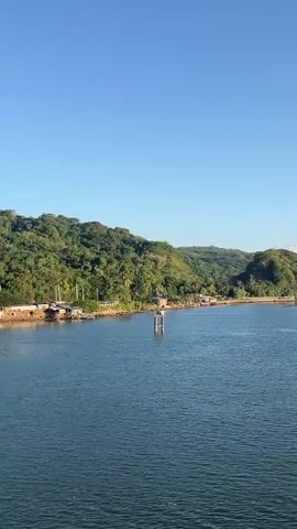 The video shows a panoramic view of a body of water with a lush, green, hilly coastline in the background. The sky is a clear, bright blue. Along the shoreline, there are several small buildings that appear to be houses or huts, some of which are built on stilts over the water. There are also some structures resembling docks or piers extending into the water. A single, tall pole or marker stands in the water in the foreground. The overall scene suggests a peaceful, tropical or coastal environment, likely during daylight hours given the brightness and clear sky. The camera appears to be stationary, panning slowly across the landscape. There are no people visible in the video.