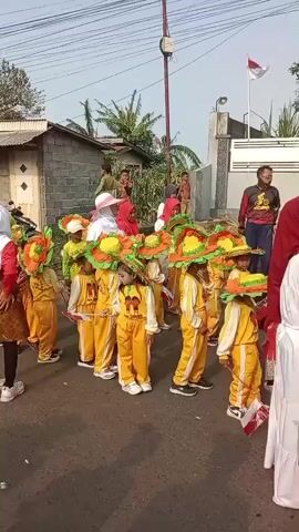 A group of young children dressed in yellow uniforms and large, colorful hats are standing in a line on a paved road. They are holding small Indonesian flags. Some adults are visible in the background, and a flag of Indonesia is flying from a pole. The scene appears to be an outdoor event or parade.