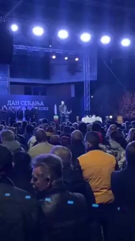 The video shows an outdoor event taking place at night. A man stands on a stage, speaking to a crowd of people. Behind him, a banner reads "Dan Secanja," and several men stand in the background. The stage is lit by bright spotlights. The camera moves to show a wall of cameras and news media equipment set up behind the crowd. The event seems to be a formal gathering, possibly a memorial or commemoration.