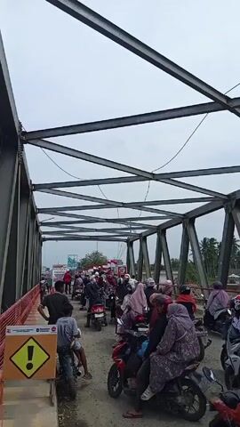 The video shows a crowd of people on motorcycles congregated on a steel truss bridge. The camera appears to be positioned on the bridge, looking down the length of the structure. The bridge has a metal frame with a lattice-like pattern of steel beams overhead. People on motorcycles are in the foreground and extend into the background. Many of the riders are wearing helmets, and some passengers are wearing headscarves. There is a yellow triangular warning sign at the front of the frame. The sky is overcast, and in the distance, trees can be seen on either side of the river the bridge spans. The road surface of the bridge appears to be gravel.