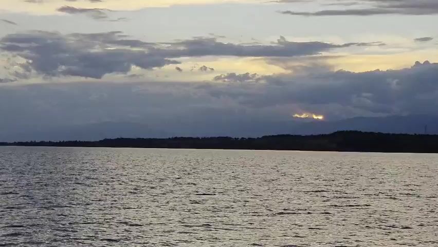 A wide shot captures a serene expanse of water under a cloudy sky, possibly a lake or a large river. The water's surface is rippled, reflecting the muted light of what appears to be either dawn or dusk. A low, dark landmass, likely a coastline with vegetation and distant mountains, stretches horizontally across the frame, dividing the water from the sky. The clouds are a mix of grey and white, with hints of yellow and orange peeking through a break in the clouds towards the right side of the image, suggesting the sun is low on the horizon and partially obscured. The overall mood is tranquil and natural, evoking a sense of peace and vastness. No people or specific objects are clearly visible, emphasizing the landscape. The scene suggests a natural, potentially remote, outdoor setting.