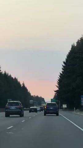 The video shows a highway scene at dusk. Several cars are driving on the highway, with trees lining both sides. A green highway sign with "EXIT 10 Olympic Dr 1/2 MILE EXIT ONLY" is visible. The sky has a pinkish hue, suggesting sunset.