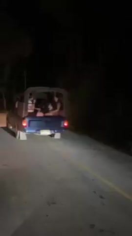 The video shows a group of motorcyclists riding on a dark road at night.  A blue pickup truck with several people in the back is parked on the side of the road. A motorcycle passes the truck. The motorcyclists drive down the road. The road is paved with yellow lines marking the lane. The environment is dark with trees lining the road.