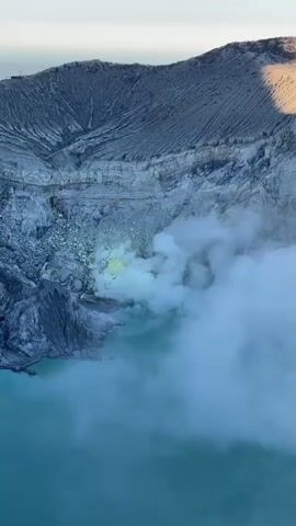 The video shows an aerial view of the crater of a volcano. The crater contains a large turquoise lake, with steam rising from a yellow vent on the side of the crater. The sides of the crater are steep and rocky, with evidence of erosion. The sky is a pale blue.