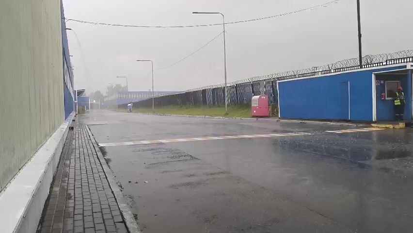 A security guard is visible inside a blue booth at the entrance to a facility. The guard is wearing a high-visibility vest and a hard hat. The facility is surrounded by a fence topped with barbed wire. The ground is wet, indicating it has recently rained or is currently raining. There are several blue shipping containers and a pink vending machine outside the booth. A speed bump is present on the wet asphalt road leading to the entrance. The overall environment appears to be an industrial or commercial area.