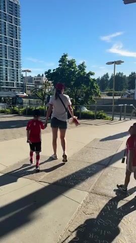In an urban environment with a tall blue skyscraper to the left and trees to the right, under a clear blue sky, a group of people are walking on a paved walkway.  A woman with blonde hair wearing a white t-shirt, denim shorts, and a red baseball cap walks away from the camera, holding hands with a young boy in a red shirt and shorts.  To their right, another woman in a pink shirt and red baseball cap walks with another young boy in a red shirt and shorts.  A person with dark hair, wearing a white and black patterned shirt, brown pants, and a backpack, walks towards the group from the foreground, facing away from the camera.  In the background, buses are visible, and streetlights are present.  The sun casts long shadows on the walkway.