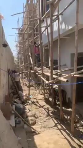 A narrow alleyway bordered by a tall, unpainted brick wall on the left and a partially constructed two-story white building on the right is filled with bamboo scaffolding. Three men are visible: one climbs the scaffolding, another is working on the ground near the wall, and the third is working on the ground beside the building. Debris and construction materials clutter the ground. The sky is clear and the sun is shining.