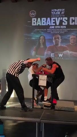 A referee in a black and white striped shirt stands to the left of an arm wrestling table, his back to the camera. He appears to be giving instructions to two arm wrestlers. On the right side of the table, an arm wrestler wearing a black sleeveless shirt and black shorts is seated. His opponent, on the left, is wearing a white t-shirt and black shorts. Both men are gripping each other’s hands on the table. The referee is positioned between them, with his hands on their backs. The table is labeled "TABLE 2" in white lettering. Behind the table, a large screen displays a poster for an event called "BABAEV'S CUP" presented by "KING OF TABLE". The poster includes the date "JUNE 13TH 2026" and a location in Switzerland. The screen also features images of two men and one woman. The arm wrestling match is taking place indoors, with a dimly lit environment and a projection screen illuminating the scene.