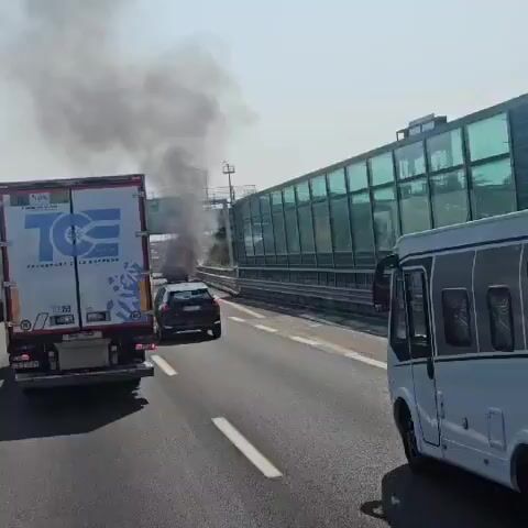 The video captures a dramatic scene on a highway, likely near Desenzano del Garda, Italy, under a bright, sunny sky. The primary event is a car fire, with thick, dark smoke billowing upwards from a vehicle engulfed in flames. The fire is intense, with orange flames visible beneath the smoke.

Several vehicles are present on the highway, all seemingly affected by the incident. In the foreground, a white recreational vehicle (RV) is driving in the right lane, partially obscuring the view of the burning car. To the left, a large white truck with the logo "TCE TRANSPORT & LOGISTICS" is visible, also in motion. Behind the burning car, a black SUV is driving away from the fire, its occupants likely concerned. Further ahead, near the fire, a tow truck with flashing yellow lights is visible, suggesting emergency services are already on the scene or en route to manage the situation.

Traffic appears to be slowed or stopped due to the obstruction. Two traffic signs are visible near the burning vehicle: one indicates a speed limit of 40, and another with a red circle and a diagonal line across it, likely prohibiting entry or a specific action. A sound barrier made of green glass runs along the right side of the highway, suggesting an urban or populated area is nearby. The overall atmosphere is one of urgency and potential danger, given the nature of the event and the presence of emergency vehicles. The time of day appears to be midday, given the harsh shadows and bright sunlight.