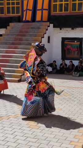 The video shows a traditional dance performance taking place in an outdoor courtyard. The main subject is a dancer wearing a vibrant, elaborate costume and a mask with a fearsome expression, possibly representing a mythical creature or deity. The costume is colorful, featuring intricate patterns and flowing fabric. The dancer is captured mid-motion, in the process of turning, with their arms outstretched. To the left and slightly behind the main dancer, another individual is visible, also in traditional attire, seated or in a bent posture. In the background, a group of spectators are seated on the ground, observing the performance. Behind them, there is a building with distinct architecture, including visible stairs with colorful treads and windows with yellow frames. The ground of the courtyard is paved with interlocking stones. The overall atmosphere suggests a cultural event or religious festival.