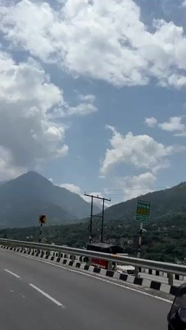 A motorcyclist, viewed from behind, rides on the right side of a highway. The motorcyclist is wearing a dark t-shirt and shorts. The motorcycle is black with a white license plate. The highway has a white line dividing the lanes and a black and white striped barrier on the right side. In the background, there are mountains covered in trees. The sky is blue with white clouds. A dark colored SUV is visible in the distance, also traveling on the highway.