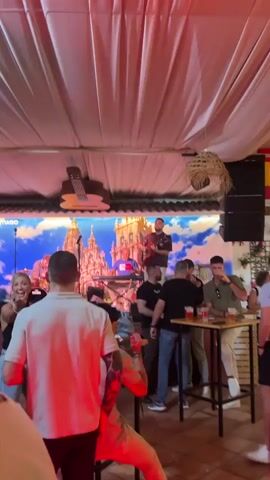 The video captures a lively evening scene at what appears to be a bar or event space in Fuengirola, Spain. A live band performs on a raised stage, featuring a male guitarist and vocalist in a maroon shirt, a male keyboardist in a purple shirt, and a drummer. Behind the band is a large backdrop depicting a grand European city or cathedral, with the word "SANTIAGO" faintly visible, suggesting Santiago de Compostela.

Numerous people, predominantly men, are gathered in front of and around the stage, socializing and enjoying the music. Many are standing, holding drinks that appear to be beer, and some are interacting at high-top tables. The venue is adorned with white drapes on the ceiling, hanging decorative elements like straw baskets and colorful pom-poms, and flags, including a Spanish flag. The space is dimly lit with warm red and orange ambient lighting, creating a festive and energetic atmosphere. The camera moves through the crowd, capturing various angles of the performance and the attendees, who seem to be in good spirits. The keyboardist's shirt also has "EL PRIMER" written on it.