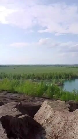 The video shows a landscape view from a high point, possibly a cliff or hill. The foreground consists of reddish-brown, rocky terrain with crevices and uneven surfaces. In the middle ground, there is a lake with turquoise-colored water, surrounded by green trees. Beyond the lake, there is a flat expanse of green fields and forests stretching to the horizon. The sky is blue with scattered white clouds. The overall impression is a contrast between the rough, rocky foreground and the serene, green landscape in the distance.