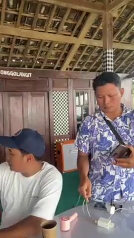 The video shows an indoor scene at a gathering of people. Two men are visible at the start, one wearing a blue baseball cap and a white T-shirt, and the other wearing a purple floral shirt, handling a charger and a portable fan on a table. The scene then pans to show several other people sitting at tables, including young children and teenagers, inside an open-air structure with a wooden roof. A woman wearing a pink hijab and floral dress is also seen, standing with her arms raised in a welcoming gesture. The location is marked with a sign that reads "PENDOPO SONGGOLANGIT."