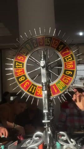 The video shows a roulette wheel surrounded by people in an indoor setting. The roulette wheel has numbers from 0 to 20, as well as 00, and is adorned with numerous small spikes around its outer edge. The wheel is mounted on a decorative silver stand. Several individuals are seen around the wheel, with some holding and stacking chips. One man, wearing a green shirt, is particularly visible on the left side of the frame. The setting appears to be a casual gambling event with individuals enjoying the game.