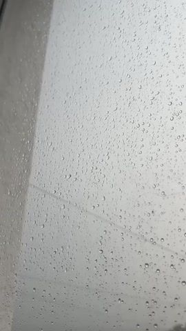 This video shows a close-up view of a window covered in raindrops. The water droplets are of various sizes and are spread across the glass, obscuring the view outside. The background is a blurry, grey sky, indicating that it is raining. There is also a faint horizontal line, possibly a wire or ledge, visible through the water droplets. The overall impression is one of a gloomy, rainy day.