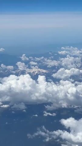 An aerial view of mountain peaks, some with snow caps, peeking through fluffy white clouds. The sky above is a clear, bright blue, fading to a lighter hue towards the horizon. The landscape below appears to be mountainous and forested. The viewpoint suggests the video was taken from an airplane.