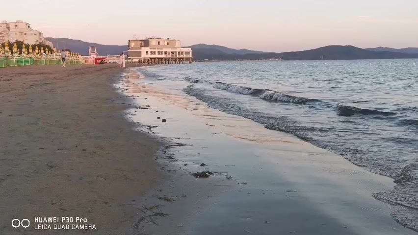 The video shows a beach with waves gently lapping the shore. In the background, there is a building on stilts over the water, with hills in the distance. The sky is a soft pink and orange, indicating either sunrise or sunset. There are some beach chairs and umbrellas on the sand. A person is walking on the beach in the distance. The video is filmed from a low angle, close to the water's edge. A watermark at the bottom of the screen indicates the video was recorded with a "HUAWEI P30 Pro LEICA QUAD CAMERA."