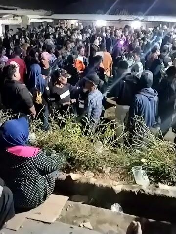 The video shows a large gathering of people outdoors at night, possibly a concert or event. The crowd appears dense, with many individuals dressed in dark clothing. Some people are wearing hats or headscarves. There are some plants and debris in the foreground, and artificial lighting illuminates the scene. Some individuals are interacting, and the overall atmosphere appears to be one of excitement or movement.