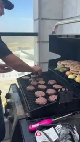 A young man with a mustache and a cap smiles and rubs his hands together near a gas grill on a balcony. The grill has many small patties cooking on one side, while the other side has grilled flatbreads, onions, and green peppers. The balcony offers a view of buildings and terrain under a sunny sky. A thermometer and meat packaging are visible near the grill.