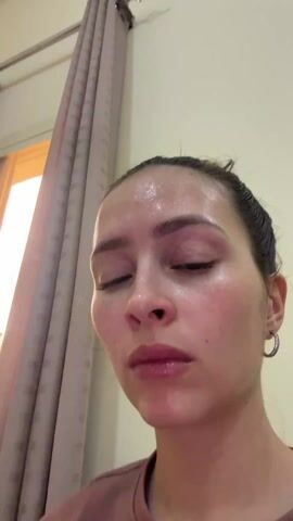 A woman with her hair in a bun holds a device up to her forehead. The device has a silver edge and emits blue light onto her forehead. She is wearing hoop earrings and a brown shirt. Her skin is shiny and appears wet. A window with a curtain is in the background.