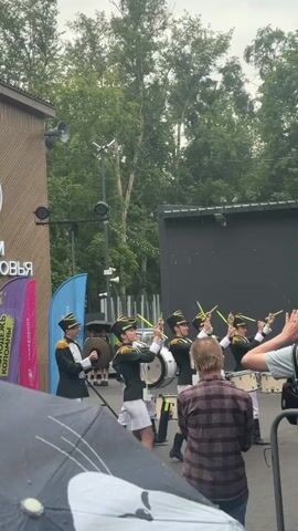 A group of young people in matching green and gold marching band uniforms, including hats, are playing instruments. Some are holding drums, while others are hitting cymbals with drumsticks or holding what appear to be tenor drums. They are standing in a line, facing away from the camera, in what looks like an outdoor area with trees and a building in the background. The sky is overcast, and some people in the foreground are holding umbrellas, suggesting it might be raining. A few spectators are visible, one of whom is filming the performance.