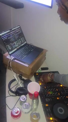 A person with multiple gold bracelets is shown standing over a table, operating DJ equipment. The table holds a laptop supported by a cardboard box, cans of Buzzballz, a glass, headphones, and a plastic cup. A TV screen is partially visible in the background. The person appears focused on the DJ equipment, with one hand on the control panel.