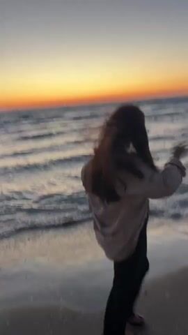 A person with long, dark hair in a light-colored top and dark pants walks left to right along a sandy beach near the water's edge at sunset. The water has small waves, and the sky is a gradient of orange, yellow, and blue. In the distance, a few rocks and a tall light pole are visible. The person has their hand near their head.