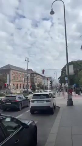 The video shows a city street scene in what appears to be Europe. Cars are lined up at a traffic light, and pedestrians are visible on the sidewalk. The camera pans upward to show the architecture of the surrounding buildings, which are large and ornate, with some greenery visible. The sky is overcast.