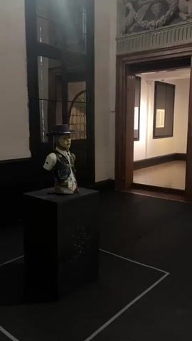 The video opens in an art gallery, displaying a stylized bust on a dark rectangular pedestal to the left. The bust depicts a figure with a light-colored face, a hat, a white shirt, and a dark vest. Behind it, a large window or glass door is visible. To the right of the bust, a dark wooden archway leads into another room with white walls and framed artwork. The floor is dark with white tape outlining a rectangular section in the foreground. The camera then slowly pans to the right, revealing a large contemporary painting on the wall. The painting features multiple figures, some appearing partially draped or nude, in various poses against a dark, reddish-brown background. A red fire extinguisher is mounted on the wall beneath the painting. As the camera continues to pan, it focuses on different sections of the painting, highlighting more of the figures and their distorted forms. The video concludes with the camera panning across the right side of the large painting.