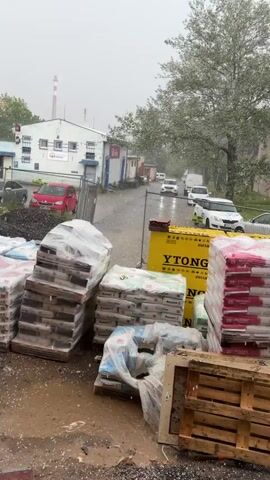The video shows a street scene during a hailstorm. The camera is positioned behind stacks of building materials, including bags of Baumit and Ytong products, and pallets. In the background, cars are driving down the wet street, and buildings are visible. The hailstones are visibly falling and accumulating on the ground, creating puddles and splashes. The weather appears to be inclement, with heavy precipitation.