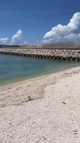 The video shows a beach on a sunny day with a blue sky and puffy white clouds. The beach is composed of small white stones, likely shells. There are several pieces of debris visible on the beach. A row of concrete tetrapods forms a barrier along one edge of the beach. The water is a clear turquoise color, and gentle waves lap the shore. The camera pans to show the sea and a distant rocky outcrop. The video ends with a view of the beach surface and the shadow of the person holding the camera.