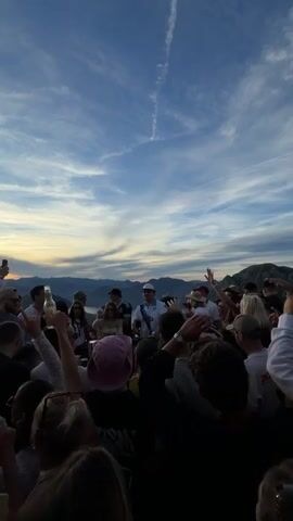 A group of people are gathered on a hilltop with a scenic view of mountains and a cloudy sky at sunset. Some individuals are raising their hands in the air, and one person in the center, wearing a white cap and a white and blue shirt, appears to be leading the group. Another person to the left is holding a bottle. The overall atmosphere suggests a celebration or an event.