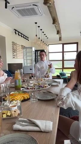 A well-lit restaurant space features a long wooden table where a group of women sit. A waiter, dressed in a white shirt and dark pants, stands behind the table, holding a bottle of wine. Various food items like cheese, oranges, and other snacks are arranged on platters across the table. There are multiple sets of cutlery, napkins, and glassware neatly laid out for each person. A vase containing white flowers serves as a centerpiece. A bottle of juice is also visible on the table. In the background, windows look out onto a green landscape. A cabinet with a glass door stands against one wall. The women at the table seem to be in conversation.