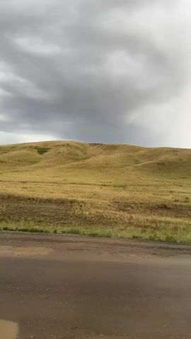The video captures a landscape scene with rolling hills covered in dry, golden grass. The sky is dramatic with heavy, dark clouds dominating the upper portion of the frame, suggesting an approaching storm or recent rain. A patch of lighter sky with some sunlight breaking through is visible on the right side. In the foreground, a wet, muddy, and uneven dirt road is present, with several puddles reflecting the sky, indicating recent rainfall. The overall mood is somber and atmospheric, with the vast, open landscape and the moody sky creating a sense of isolation. There are no people or specific objects visible apart from the natural elements of the landscape and the road. The time of day is difficult to pinpoint due to the overcast conditions, but the light suggests it could be late afternoon or early evening. The location context of Karagayl, Kazakhstan, implies a semi-arid or steppe environment, which aligns with the visual depiction of dry grasses and rolling hills. There is no visible text in the media file.