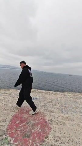 A young man dressed in a black hoodie and black sweatpants is walking along the edge of a concrete structure. The concrete surface has some colorful paint markings on it. Behind him is a body of water that appears to be the sea or a large lake, under a cloudy sky. In the distance, there is a hint of land. The man is walking from right to left in the frame.