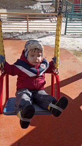 A young child is on a swing set. The child wears a red and white jacket, gray pants, black and yellow boots, gloves, and a hat. The swing has a red frame, a blue seat, and yellow ropes held by chains. The ground under the swing is orange. The child is sitting on the swing and looks straight ahead. A shadow of the child and the swing set is cast on the ground. Behind the swing set, there is a metal fence, some patches of snow, and buildings. There are a few people walking in the background.
