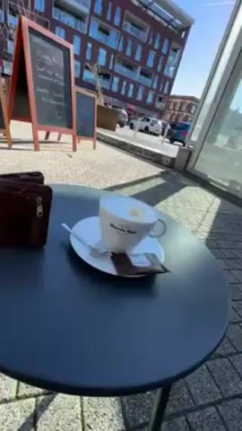 The video shows a cup of cappuccino on a black table, with a small chocolate cookie and spoon next to it on the saucer. The cup is white and has the words "Blanche Dael Maastricht Anno 1878" printed on it. The background is a street scene with buildings, cars, and a chalkboard menu. The camera zooms in on the cup of cappuccino.