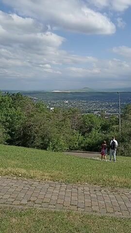 This media file depicts a scenic overlook in Pyatigorsk, Russia. The foreground features a cobblestone path and a lush green grassy slope, suggesting a park or natural area. Beyond this, a dense band of trees forms a visual barrier before opening up to reveal a sprawling cityscape in the middle ground. In the distance, a hazy horizon line meets a bright, cloudy sky, indicative of a daytime scene.

Two individuals, a woman and a man, are walking down a paved path that winds through the trees. The woman, in a red top and blue shorts, carries a backpack. The man, in a white shirt and dark pants, also has a backpack. They appear to be enjoying a leisurely walk, their posture suggesting a relaxed mood.

The overall setting is that of a picturesque vantage point offering panoramic views of the city and its surrounding landscape. The bright daylight and the presence of fluffy white clouds suggest pleasant weather conditions. The city's layout, visible in the mid-ground, includes numerous buildings and hints at a moderately sized urban area. The hazy distance and the overall composition evoke a sense of calm and appreciation for the natural and urban environment. No visible text is present in the media file.