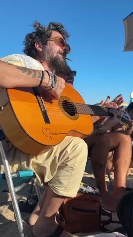 A man with curly dark hair, a beard, and sunglasses is playing an acoustic guitar on a beach. He is wearing a light-colored t-shirt and light-colored shorts. Another man with a shaved head, sunglasses, and a dark shirt is sitting next to him, singing and clapping his hands. Both men are sitting in beach chairs. The guitar has a heart drawn on it with writing inside. The background is a clear blue sky and a beach. Other people are visible in the background, along with beach umbrellas. A brown bag is on the sand in front of the man playing the guitar.