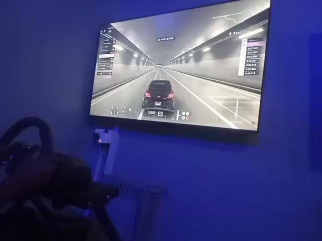 A person is playing a racing video game on a simulator. The game is displayed on a large TV mounted on a blue wall. The person is holding a steering wheel attached to a rig. The game shows a black car driving through a tunnel. A list of lap times is visible on the right side of the screen. A white radiator is visible under the TV. The room appears to be dimly lit.
