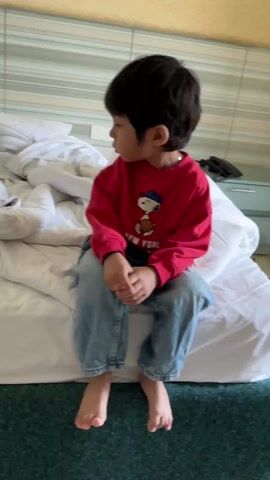 A young boy sits on a made bed, looking to his left and then toward the camera. The boy has dark hair and is wearing a red sweater with a cartoon character printed on it, and light blue jeans. He is barefoot. The bed is covered with a white comforter. The bed is against a wall with a striped, light blue pattern. A small white bedside table with a black phone is next to the bed.